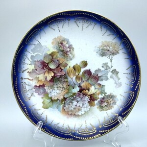 May include: A round porcelain plate featuring a central botanical illustration of green gooseberries and clusters of white and purple hydrangea flowers. The plate has a deep cobalt blue border with gold filigree accents along the outer rim.