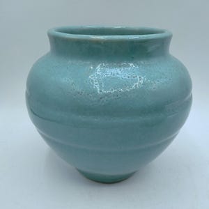 May include: A light blue ceramic vase with a rounded body and a slightly flared rim. The vase has a textured surface and horizontal ridges around the middle. The overall shape is reminiscent of a classic urn, suitable for floral arrangements or decorative display.