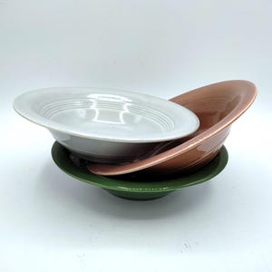 May include: A stack of three ceramic bowls in varying colors. The top bowl is white, the middle bowl is a muted terracotta orange, and the bottom bowl is a deep forest green. Each bowl features a wide, flat rim and concentric circular ridges.