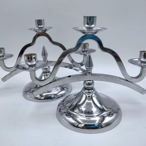 May include: Two silver-colored candelabras with a curved design. Each candelabra has a round base, a central spike, and three candle holders. The polished metal reflects light, creating a shiny appearance.