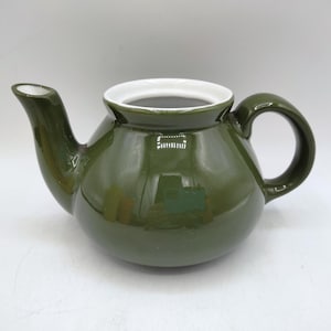 May include: A green ceramic teapot with a rounded body, a curved spout, and a large, arched handle. The interior rim is white, contrasting with the dark green exterior. The teapot is designed for brewing tea.