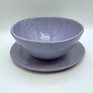 May include: A handmade, lavender-colored ceramic bowl and saucer set. The bowl has a textured interior and a rounded shape, sitting atop a matching, flat saucer. The set is a soft, muted purple, perfect for serving food or as a decorative piece.
