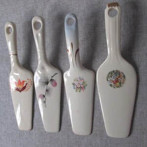 May include: A set of four off-white ceramic cake servers with varying floral and botanical motifs. Each server features a long handle with a circular hole at the top, a flat blade, and unique decorative patterns including tulips, pine cones, and floral bouquets.