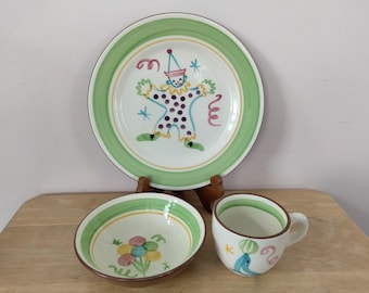 May include: A set of three ceramic dishes with a whimsical clown and floral design. The set includes a large plate, a bowl, and a mug. The plate features a clown with a party hat, the bowl has a floral design, and the mug has a blue fish.