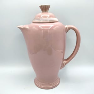 May include: A pink ceramic coffee pot with a white lid and a white trim around the lid. The pot has a rounded body and a handle on the side.