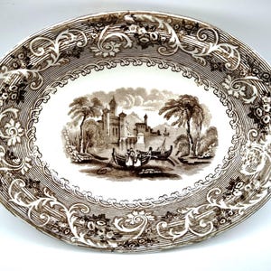 May include: Oval ceramic serving platter with a brown transferware design. The center features a landscape with a castle, boats, and figures. The border has floral and striped patterns.