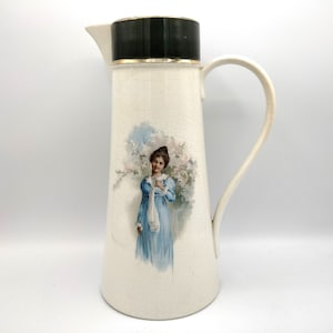 May include: Cream-colored ceramic pitcher with a black top and gold trim. A painted illustration of a woman in a blue dress is on the front. The pitcher has a curved handle and a spout.
