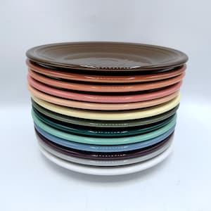 May include: A stack of colorful ceramic plates in various shades, including brown, orange, pink, yellow, green, blue, and white. The plates are round and appear to be new.
