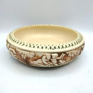 May include: Cream-colored ceramic bowl with a detailed relief design. The rim features a green and white pattern, while the sides depict figures and foliage in brown tones. A decorative piece for display or serving.