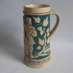 May include: A tall ceramic beer stein featuring a deep teal body with cream-colored relief carvings of a figure standing next to a barrel and hop vines. The base includes the text "Wundertropf" in black lettering, with a matching cream handle.