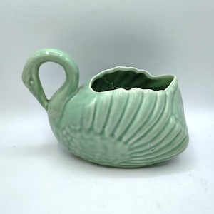 May include: A light green ceramic swan-shaped planter. The planter features a curved neck handle and a body with textured, stylized feathers. The interior is a darker green. The swan is a decorative item.