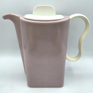 May include: A mauve-colored ceramic teapot with a white lid and handle. The teapot has a rectangular body with a curved spout and handle. The handle is a curved, off-white color.