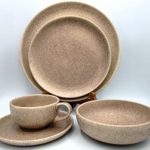 May include: A speckled, tan-colored ceramic dish set. The set includes two plates, a bowl, a teacup, and a saucer. The plates are stacked behind the other pieces. The bowl is oval-shaped. The teacup has a handle.