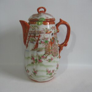 May include: A tall, antique porcelain teapot with a lid, featuring a hand-painted scene of figures and a cart. The teapot has a white base with a detailed design, accented with orange and gold trim. The handle and spout are orange.