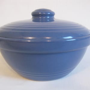 May include: A blue ceramic bowl with a matching lid. The bowl has a rounded shape with a slightly flared rim and a series of horizontal grooves around the base. The lid has a knob handle.