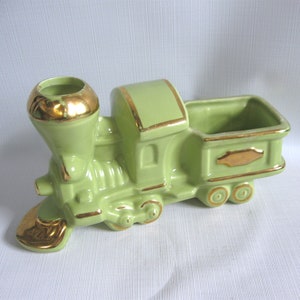 May include: A ceramic train planter in a light green color with gold accents. The train features a smokestack, a compartment, and wheels. The gold trim is on the smokestack, the front, and the compartment. The planter is designed to hold small plants or flowers.
