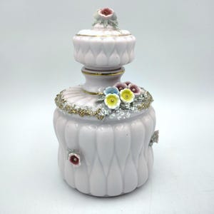 May include: A light pink porcelain decorative jar with a lid, adorned with small, colorful flowers and gold accents. The jar has a fluted design and additional floral embellishments. A delicate piece for display.