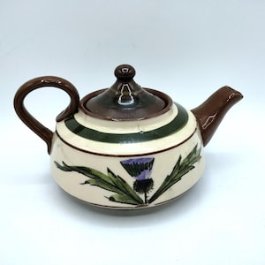 May include: A ceramic teapot with a cream-colored body, a dark brown handle, spout, and lid. The teapot features a horizontal dark green band around the upper section and a hand-painted purple and green thistle floral design on the front.