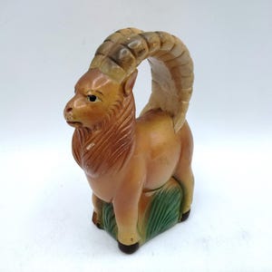 May include: A ceramic figurine of a mountain goat, featuring large curved horns and a detailed mane. The goat is painted in shades of brown and tan, with green accents at the base, creating a rustic aesthetic. The figurine is a decorative item.