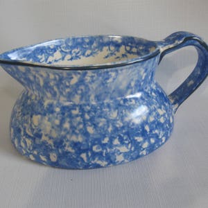 May include: A blue and white speckled ceramic pitcher with a curved handle and a spout. The pitcher has a rounded body and a black rim around the top. The handle is also blue and white speckled.