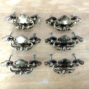 May include: A set of six brass-toned metal drawer pulls featuring a decorative, ornate backplate with a curved, swinging handle. Each pull is secured with two metal bolts extending from the back, showcasing a polished, reflective finish.
