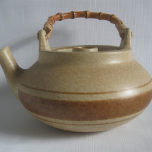 May include: A tan ceramic teapot with a bamboo handle. The teapot has a rounded body with a wide, dark brown band around the middle and a spout on the side. The handle is made of bamboo and woven material.