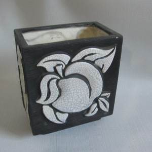 May include: Black square ceramic planter with a white floral design. The planter has a matte black exterior and a white interior. The floral design is raised and has a cracked, textured appearance. The planter is suitable for indoor or outdoor use.