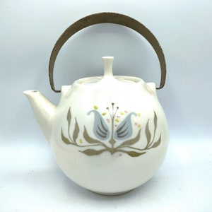 May include: Cream-colored ceramic teapot with a metal handle. The teapot features a floral design in gray, blue, and yellow. The spout and lid are visible. A vintage kitchenware item.