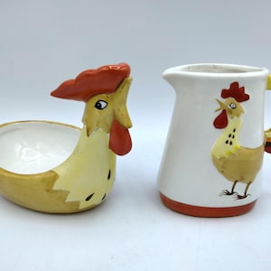 May include: A ceramic rooster-shaped dish and a ceramic pitcher with a rooster design. Both pieces are white with brown, yellow, and orange accents.