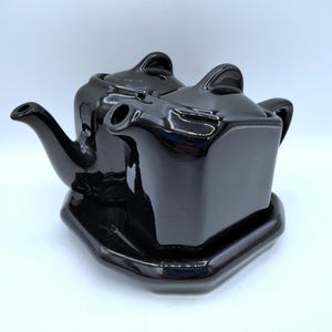 May include: A black ceramic teapot set with a square design. The set includes a teapot with a lid and a matching base. The teapot has a curved spout and a handle on top of the lid. The set is glossy and reflects light.