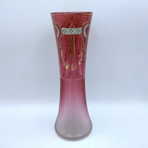 May include: A tall, elegant vase with a flared base and a slender neck. The glass transitions from a clear base to a gradient of pink, with a deep red top. Gold and white decorative elements adorn the upper portion of the vase.