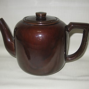 May include: A brown ceramic teapot with a lid and a curved spout. The teapot has a handle and a glossy finish. The design is simple and classic, suitable for serving tea. The teapot is a warm, earthy brown color.