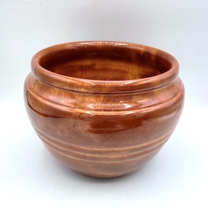 May include: A glazed, brown ceramic planter with a wide, open top and a rounded body. The planter has horizontal ridges around the exterior and a glossy finish. The interior is a darker shade of brown. This decorative pot is suitable for indoor or outdoor use.