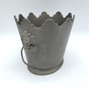 May include: A matte gray metal bucket with a scalloped top edge and a circular base. The exterior features a decorative lion head emblem on the side, which holds a dangling metal ring handle. The surface has a weathered, metallic finish.