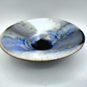 May include: A ceramic bowl with a unique design. The bowl features a blend of colors, including white, blue, and gray, creating a marbled effect. The center is a dark, glossy black. The rim is a light brown.