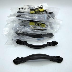 May include: A collection of matte black cabinet handles with ornate, curved ends. Several handles are individually wrapped in clear plastic packaging with yellow labels, while two identical handles are unwrapped and displayed in the foreground.