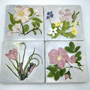 May include: Set of four square ceramic tiles with pressed flower designs. Each tile features a different flower arrangement with pink, white, and yellow flowers on a light blue background.
