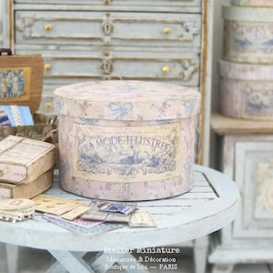 May include: A collection of miniature vintage-style boxes and containers. The central piece is a large, round box with a floral pattern and a label that reads "LA MODE ILLUSTREE." Other items include small rectangular boxes, a woven basket, and various ephemera.