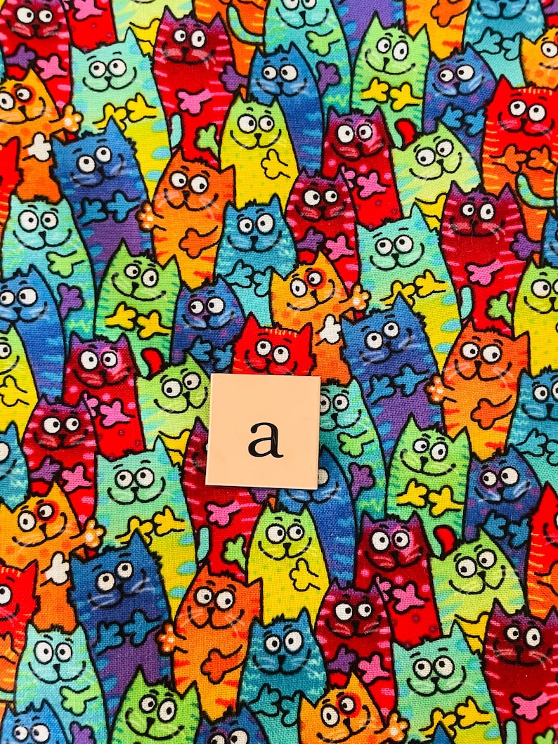 Pode incluir: Um tecido colorido com um padr&atilde;o repetitivo de gatos de desenho animado em v&aacute;rias cores e poses. A letra "a" est&aacute; impressa em um quadrado branco.