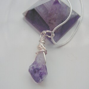 Puede incluir: Un collar de cadena de plata con un colgante de amatista púrpura. El colgante es un cristal grande y facetado con un cristal de amatista más pequeño y áspero colgando de él.