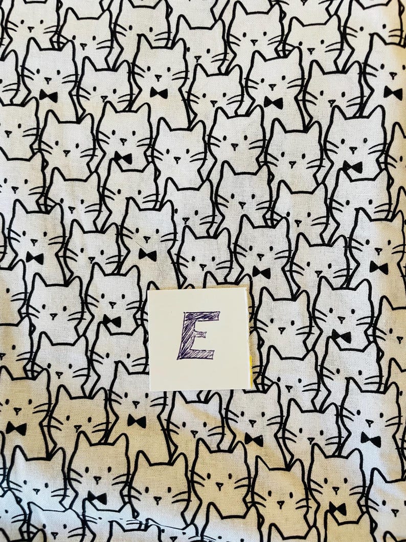 Pode incluir: Tecido branco com um padr&atilde;o repetido em preto e branco de gatos usando gravatas borboleta. A letra "E" est&aacute; escrita em roxo em um cart&atilde;o branco.