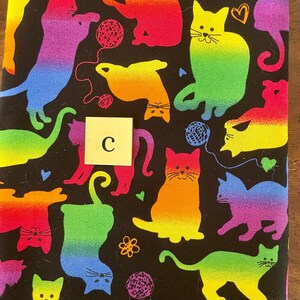 Pode incluir: Um tecido preto com um padr&atilde;o de arco-&iacute;ris de gatos brincando com fio. O tecido tem uma letra "C" branca no centro.