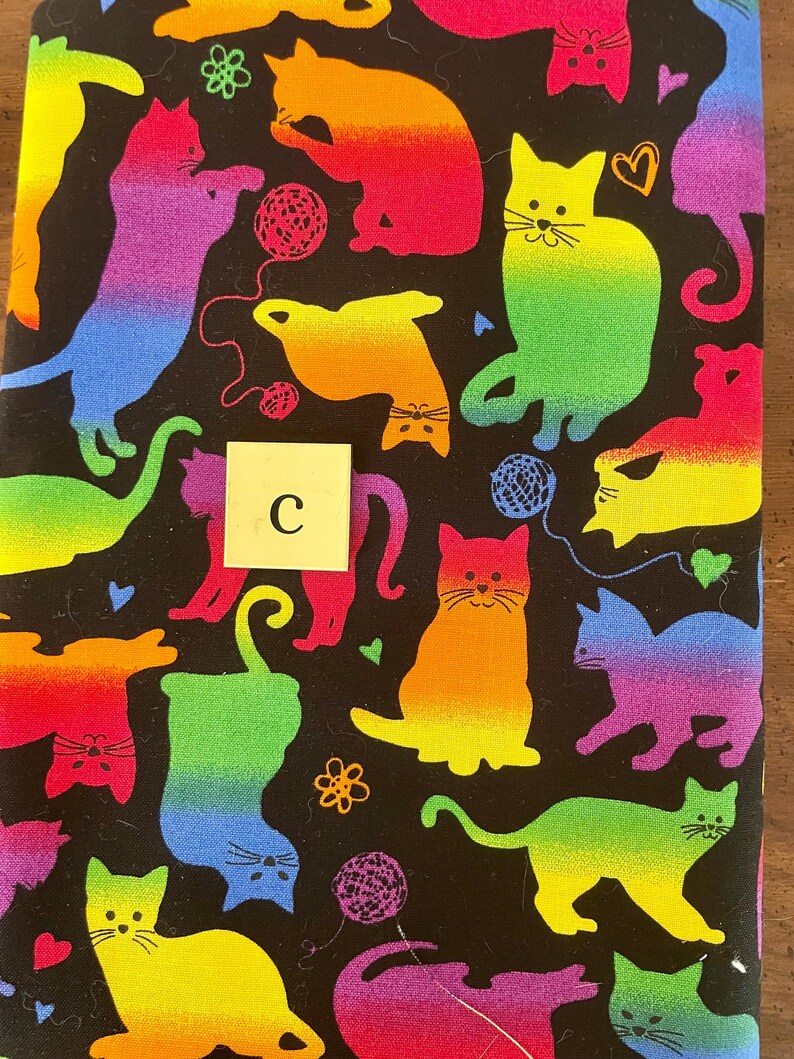 Pode incluir: Um tecido preto com um padr&atilde;o de arco-&iacute;ris de gatos brincando com fio. O tecido tem uma letra "C" branca no centro.