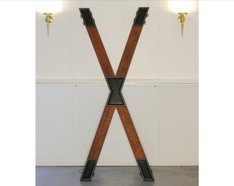 May include: A wooden X-shaped frame with black leather padding and metal accents. The frame is standing upright against a white wall.