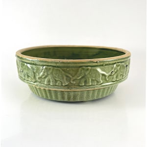 May include: A green ceramic bowl with an embossed elephant design around the rim. The bowl has a wide, shallow shape and a slightly textured surface. The rim is a lighter shade of green.