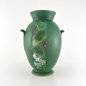 May include: A green ceramic vase with a flared rim and two small side handles. The body features a central vertical panel decorated with raised relief carvings of green leaves, brown branches, and small white flower clusters.