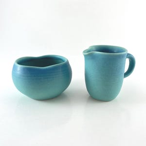 May include: A ceramic sugar bowl and a matching creamer pitcher, both featuring a matte turquoise blue glaze with subtle color variations. The sugar bowl has a rounded, organic shape with a wavy rim, while the creamer has a spout and handle.
