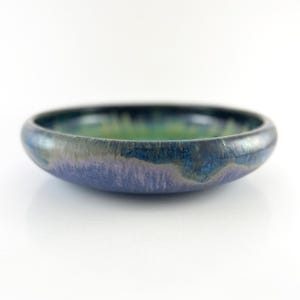 May include: A small, round ceramic bowl with a low profile. The bowl features a gradient of colors, including blue, green, and purple. The rim is dark blue, and the interior has a green hue. The bowl is likely handmade and has a glossy finish.