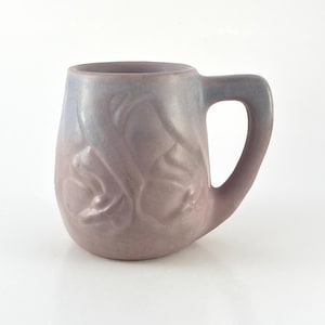 May include: A ceramic mug with a handle, featuring a gradient of colors from lavender to pale pink. The mug has a textured surface with a raised, organic design. The mug is designed for hot beverages.