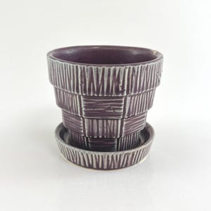 May include: A deep purple ceramic plant pot with a matching saucer, featuring a tiered, stepped silhouette and a textured surface pattern of vertical lines and horizontal bands that create a woven, basket-like appearance with a glossy finish.